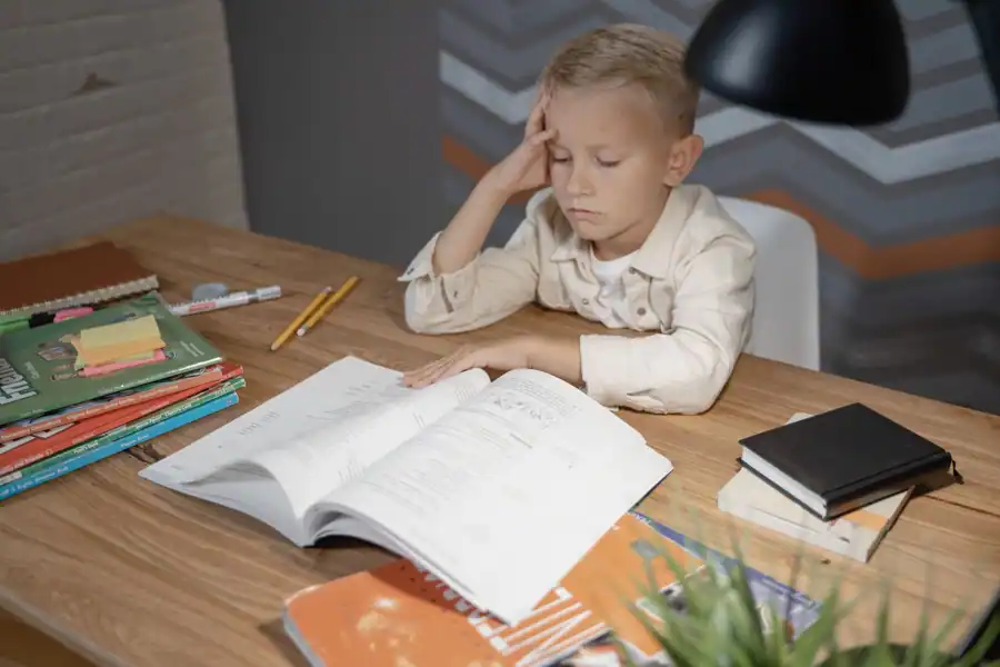 A young boy appears deep in thought while studying at his desk filled with books and notebooks.