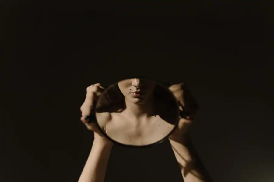 Artistic portrait of a woman holding a mirror, reflecting her face, with a dark, moody background.