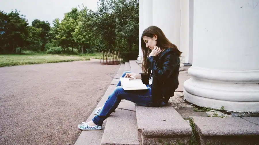 A woman reads a book while seated on stone steps outside, surrounded by nature.